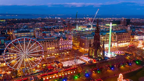 Aerial view of the Edinburgh Christmas Market and Winter Wonderland with a Ferris wheel, rides, and city lights at dusk.
